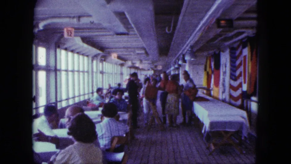 Passengers in 1960s attire occupy a long shipboard dining space, with tables by the windows and flags along the opposite wall.