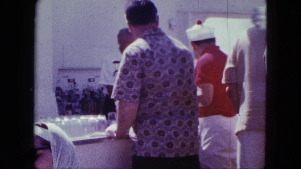 1963 shipboard buffet frame showing travelers queued at a counter with bottles and serving containers, filmed from a close handheld viewpoint.
