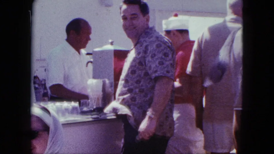 In this 1963 San Diego frame, a customer turns toward the home-movie camera while patrons and a bartender remain active at the counter.
