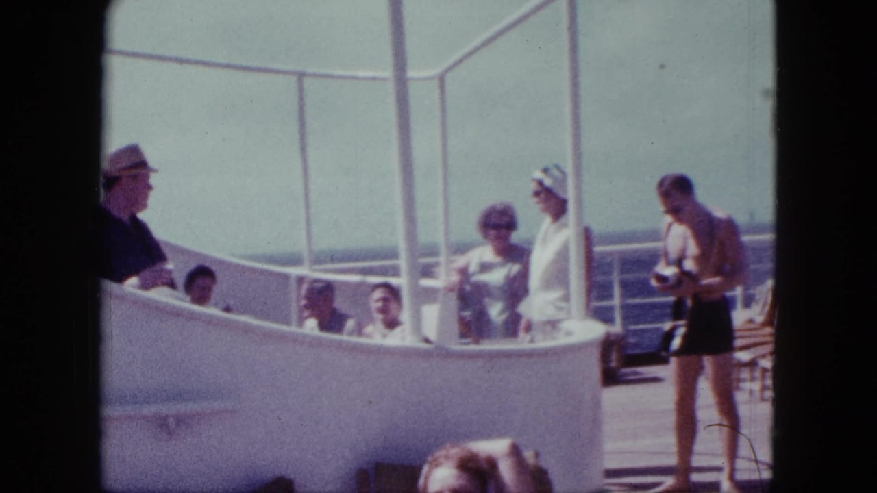 Passengers stand and sit on a cruise ship deck near white railings and deck chairs.