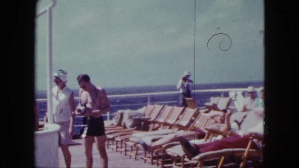 1963 cruise ship deck scene with rows of wooden lounge chairs, sunbathers, and a standing passenger holding a camera near the railing.