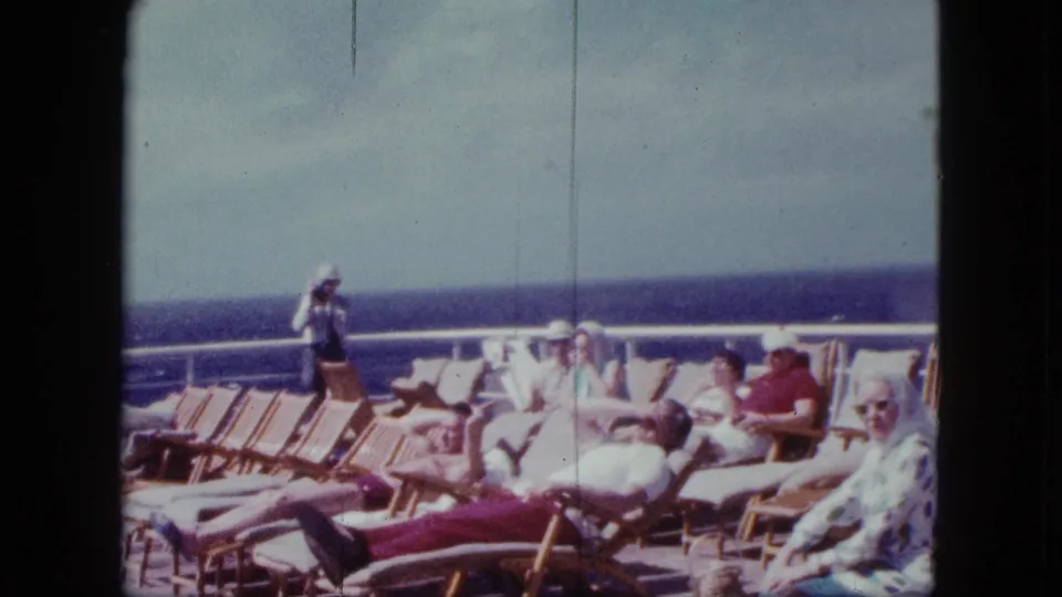 1963 cruise ship deck view of passengers reclining in wooden chairs under a bright sky, with the ocean horizon in the background.