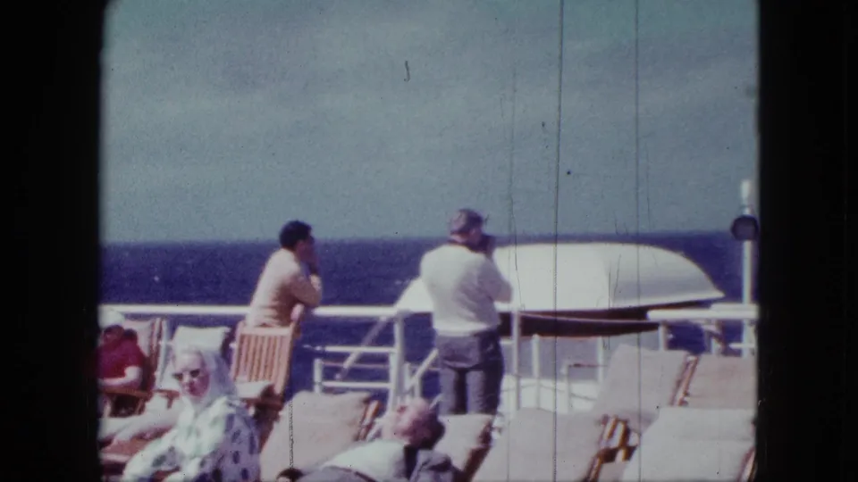 1963 8mm cruise-liner deck frame with a seated woman, standing adults at the rail, deck chairs, and ocean background.