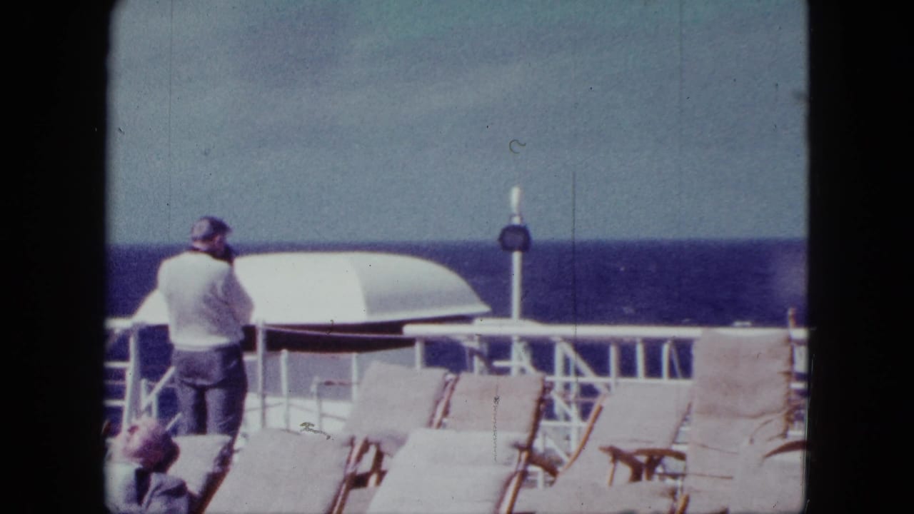 Passenger stands by a ship railing near deck chairs with the ocean horizon beyond.