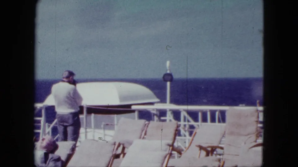 1963 home-movie frame of a cruise ship deck near San Diego, with a passenger at the rail, lounge chairs, and open ocean beyond.