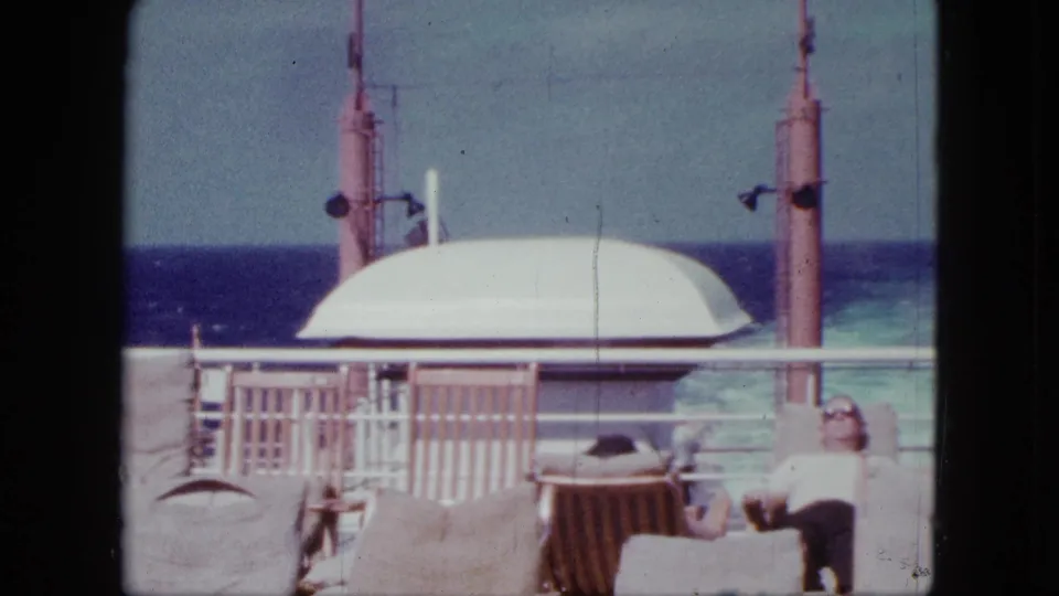 A seated passenger appears on a 1963 cruise ship deck near lounge chairs, railings, a curved canopy, and the ocean off San Diego.