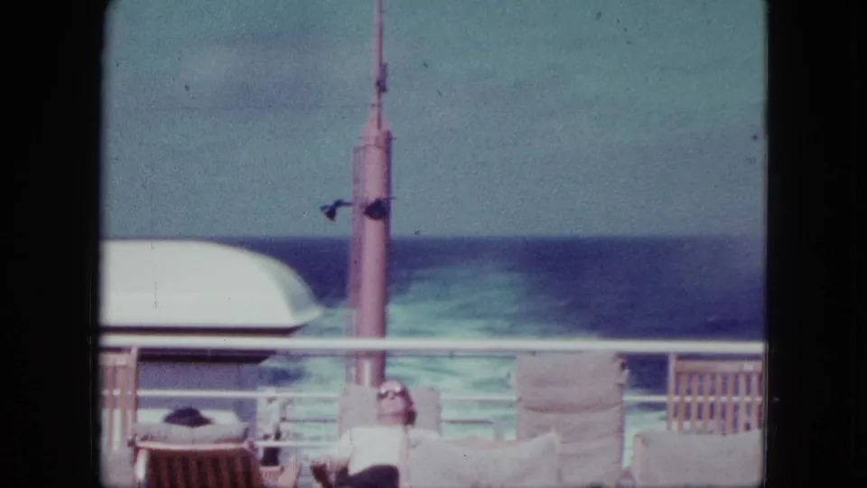 Home-movie view of a ship deck in 1963, showing a reclining passenger low in frame beneath the railing with ocean and deck equipment beyond.
