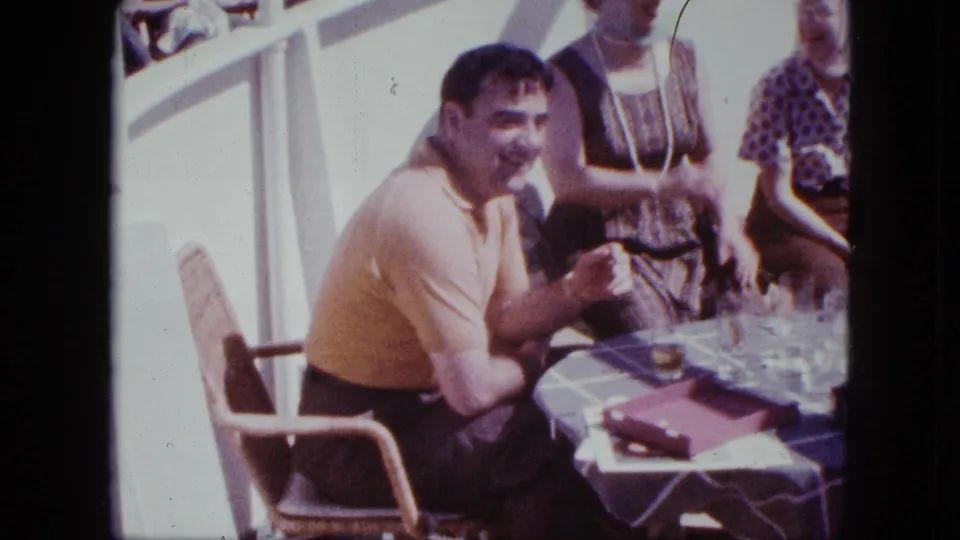 1963 shipboard home-movie frame showing adults gathered at a deck table, with a man smiling toward the camera and tableware in front of him.