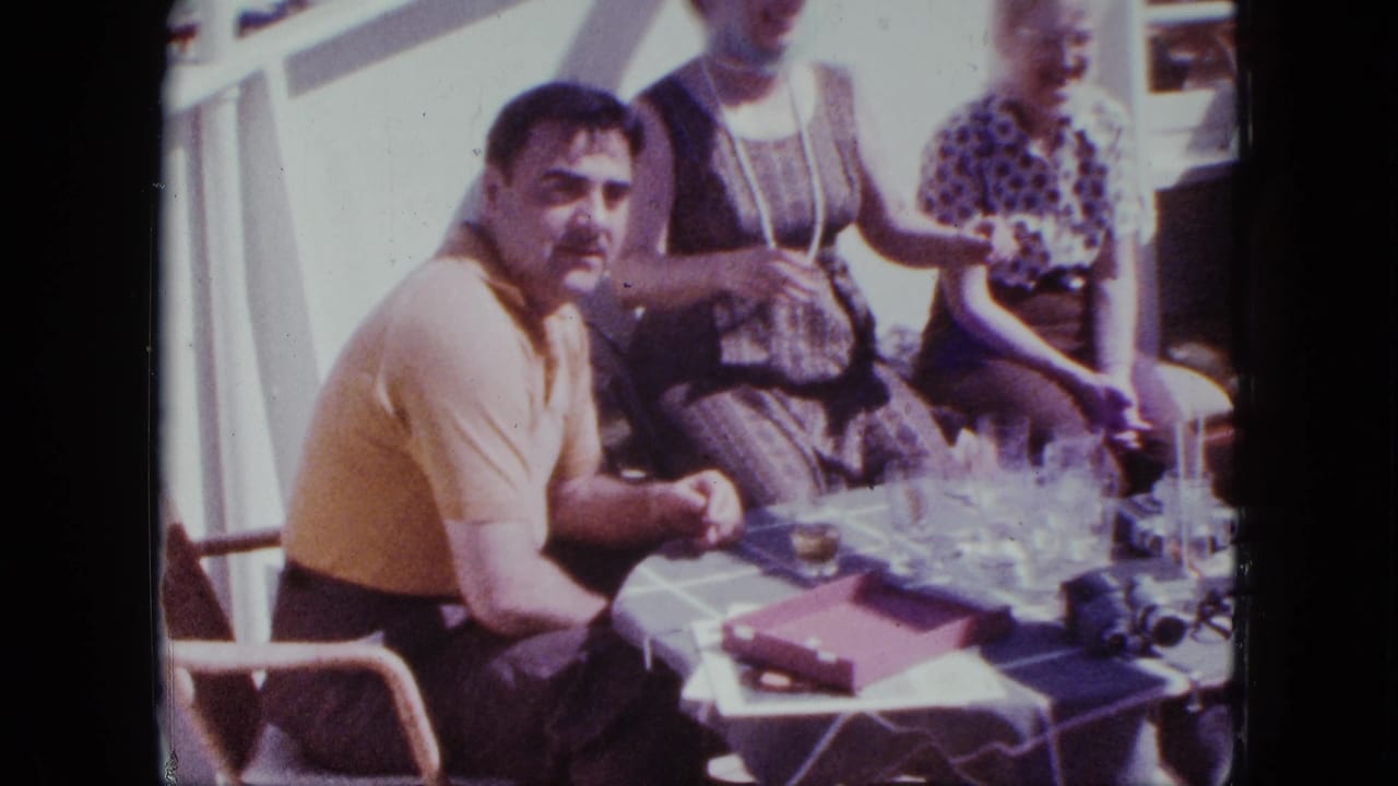 Man in yellow shirt turns toward the camera beside seated women and table glasses.