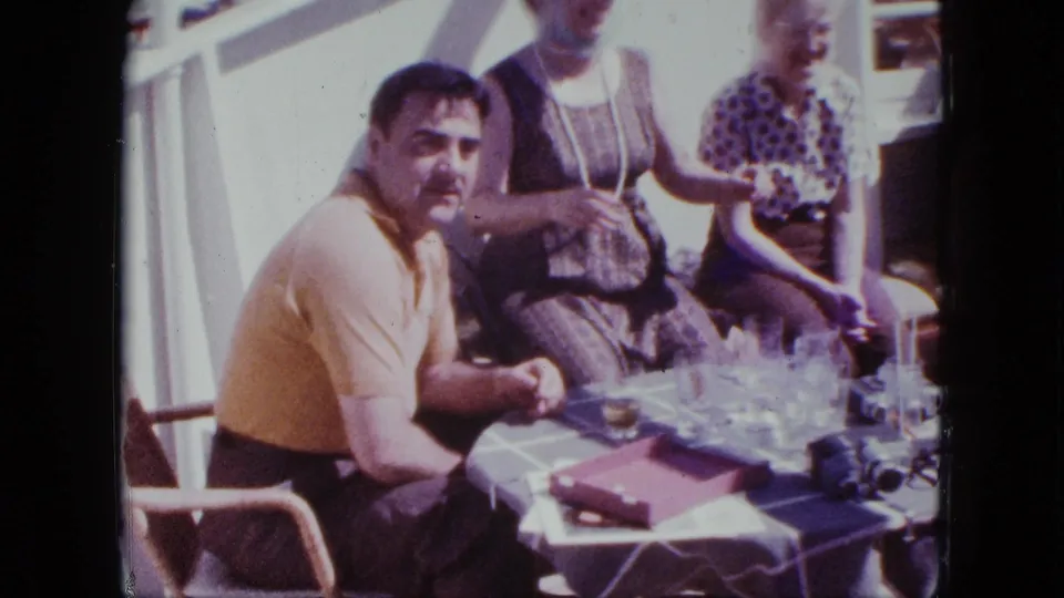 1963 San Diego home-movie frame with a man in a yellow polo facing the camera at a deck table with seated women.