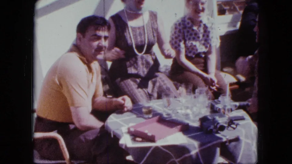 1963 San Diego home-movie frame showing adults around a checkered table with glasses, a red box, and a small camera.