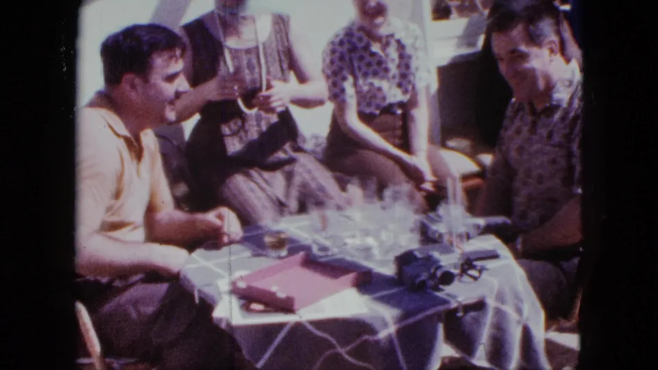 1963 San Diego home-movie frame of adults seated on a deck as a man at right joins the table conversation.