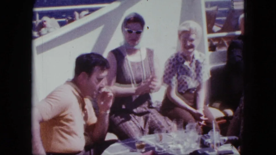 1963 San Diego home-movie frame of adults socializing on a boat deck, with sunglasses, patterned clothing, and drinks visible.