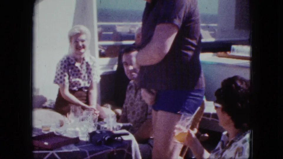 1963 San Diego boat outing frame with a standing man, seated adults, glasses, and a camera on the table.