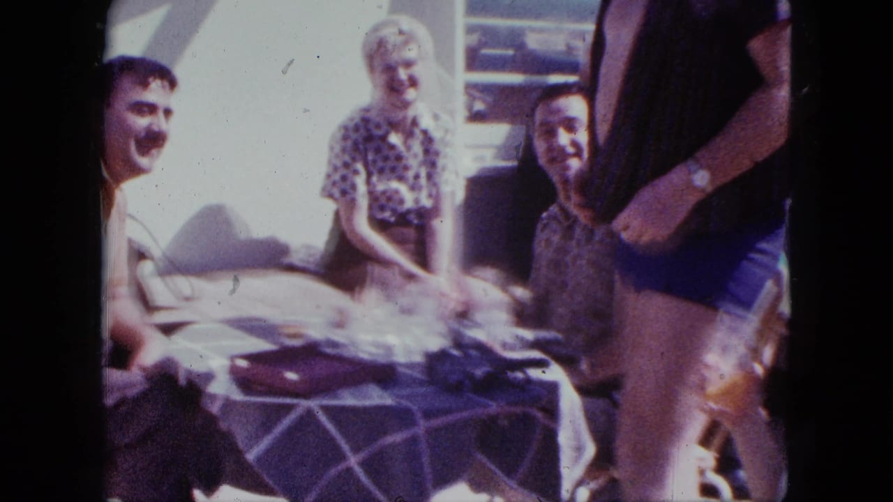 Adults gather at a deck table, including a smiling woman and seated men in bright sun.