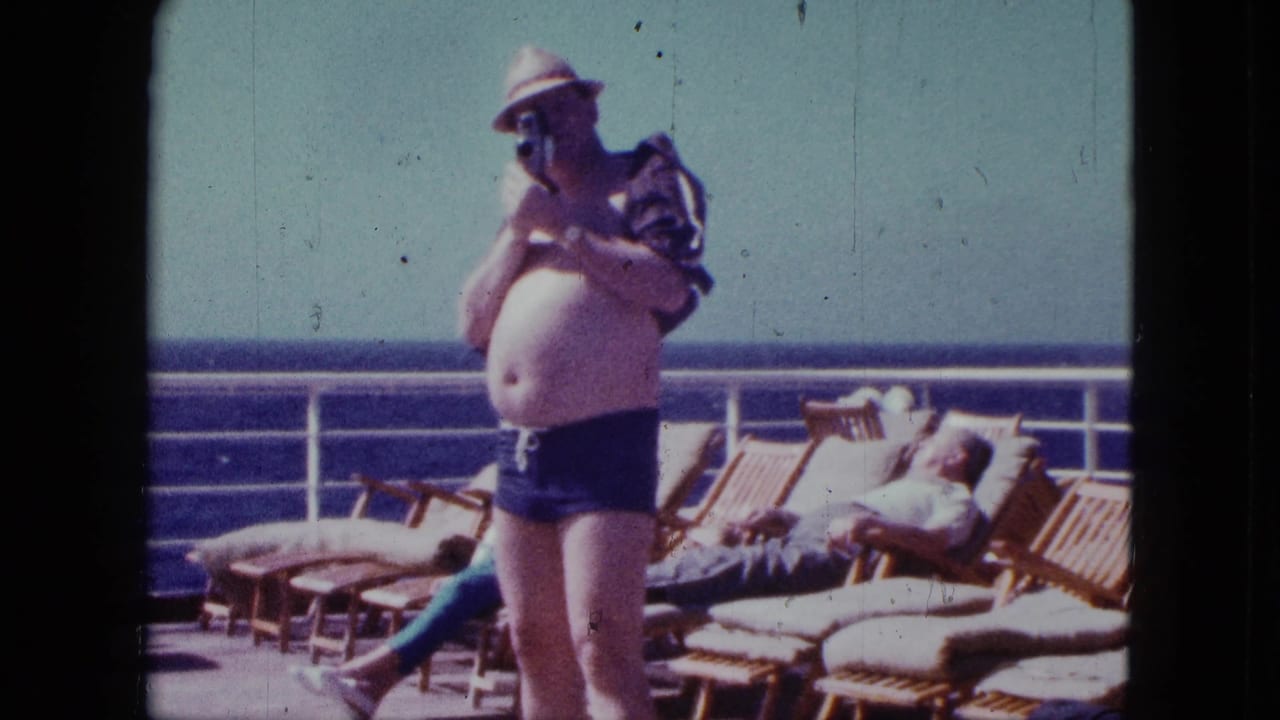 Man in straw hat and swim trunks holds a camera on a ship deck near lounge chairs.