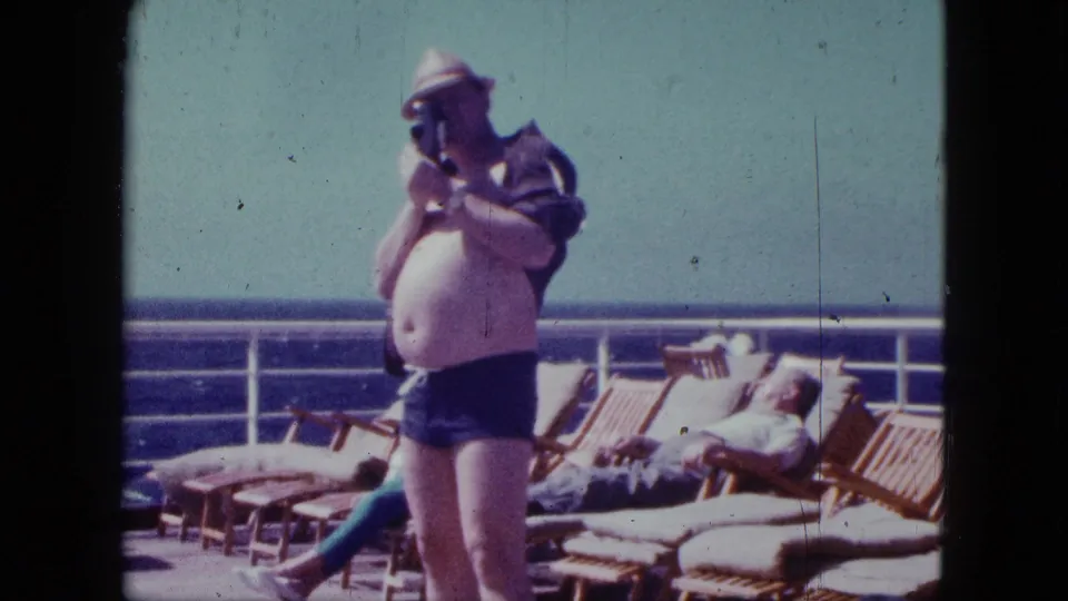 1963 cruise footage shows the man with a camera centered on deck, with wooden loungers, a reclining passenger, railing, sky, and sea visible.