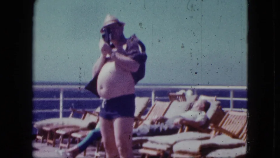 A 1963 home-movie frame shows the cruise-deck photographer looking through his camera, with lounge chairs, a reclining adult, railing, and sea behind him.