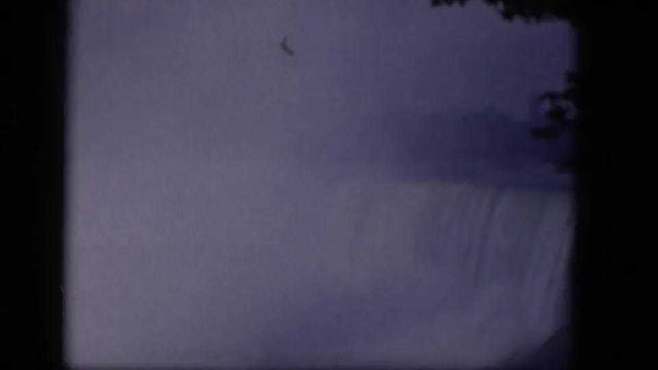 1964 8mm frame of a mist-covered waterfall, with dark gate edges, soft focus, and faint tree silhouettes near the right side.