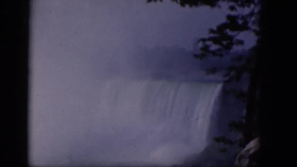 Blue-tinted 1964 8mm view of mist rising from a waterfall, with dark frame borders and tree branches silhouetted at the right.