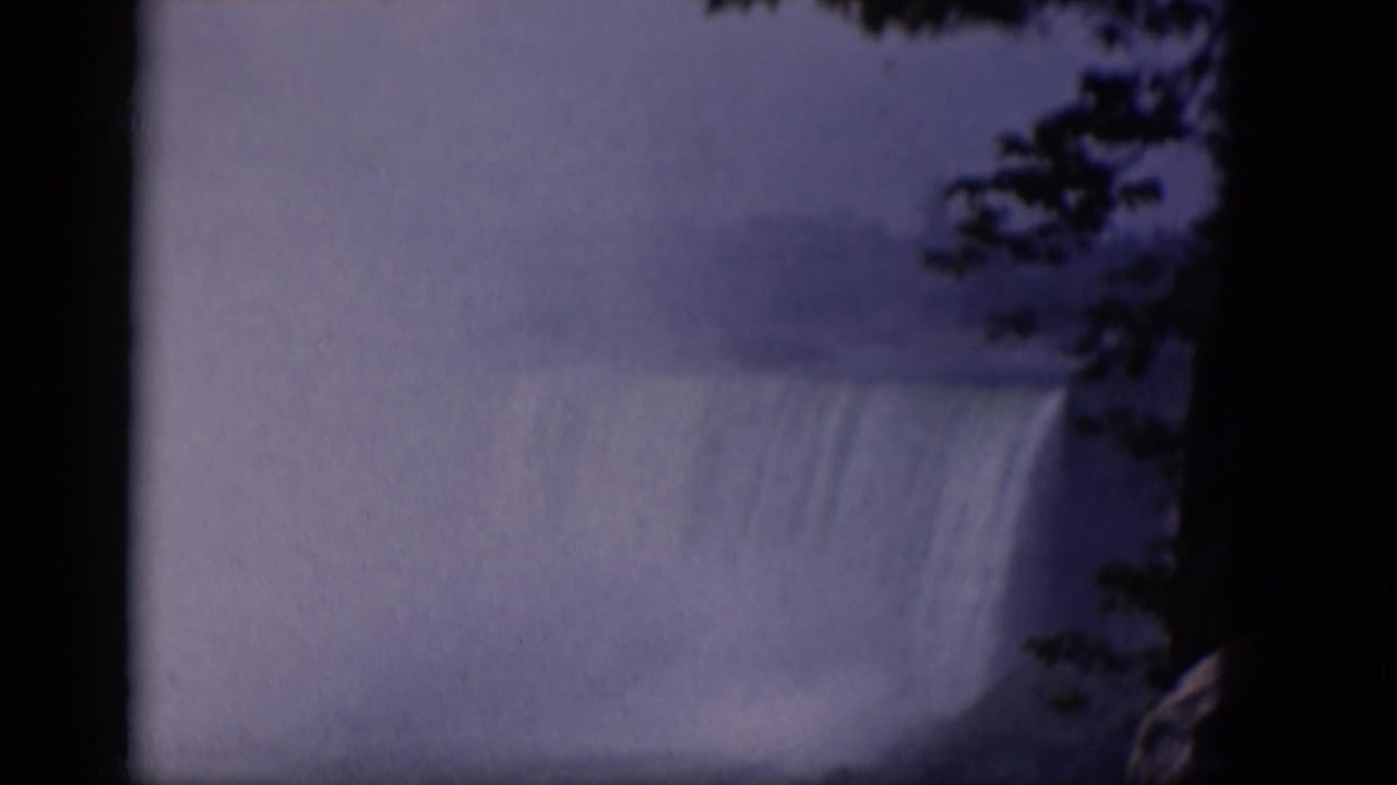 1964 8mm home-movie frame of a large misty waterfall, with dark tree branches and shadowed film edges framing the cascade.