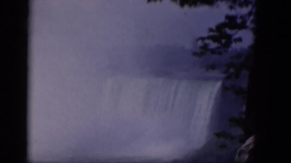 1964 8mm home-movie frame of a large misty waterfall, with dark tree branches and shadowed film edges framing the cascade.