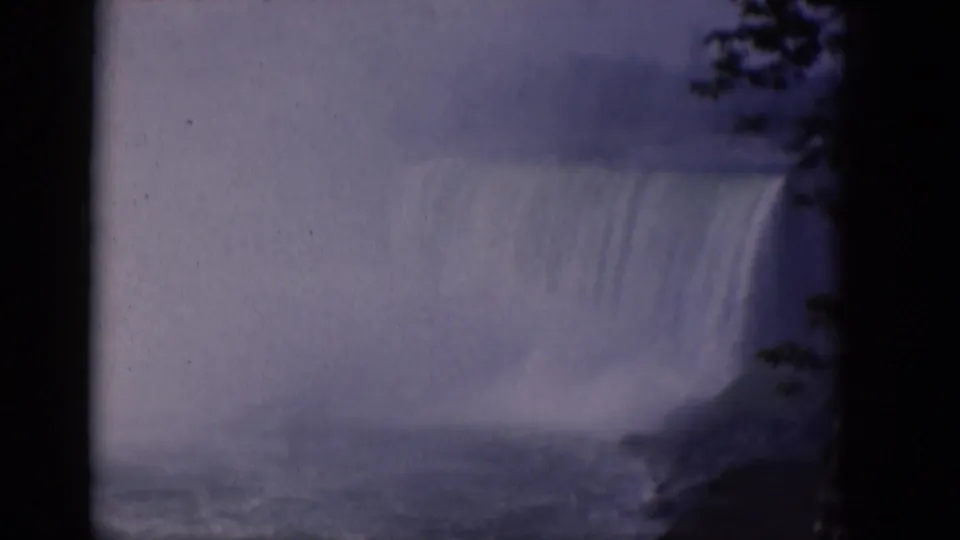 1964 8mm travel frame showing a broad waterfall and spray, with rough water below and dark foliage along the right edge.