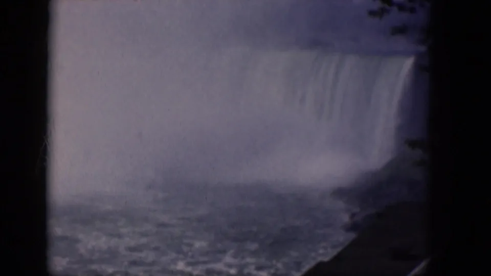 1964 8mm frame of a mist-covered waterfall viewed through shadowed trees, with bright spray and rough water near the base.