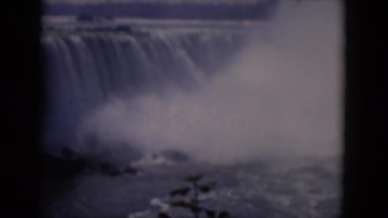 Soft 8mm frame of a large waterfall dropping into mist behind dark film borders.