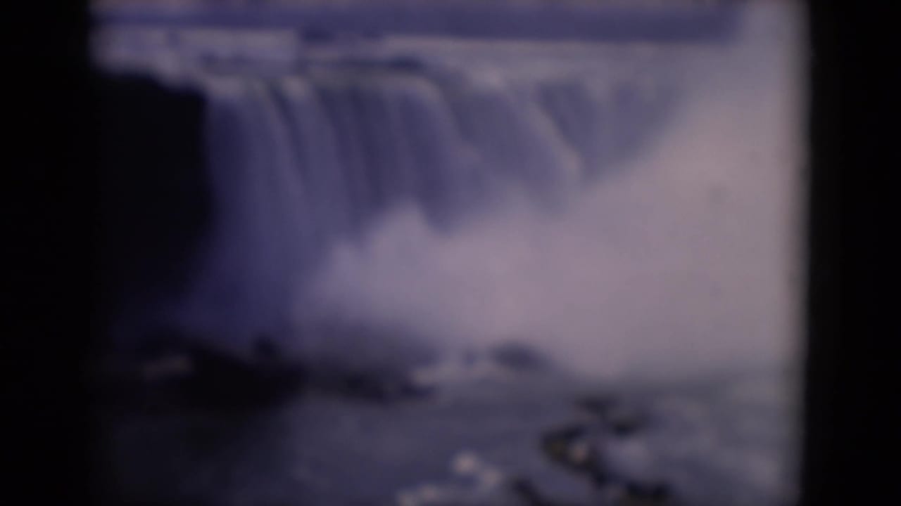 1964 8mm travel footage frame with a broad waterfall lip, heavy mist, dark cliff shape, and distant treeline.
