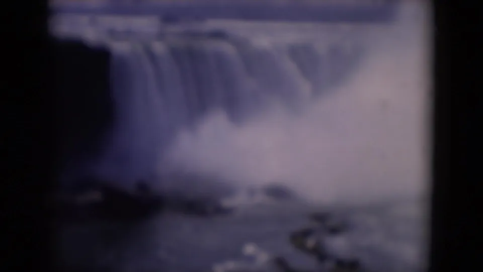 1964 8mm travel footage frame with a broad waterfall lip, heavy mist, dark cliff shape, and distant treeline.