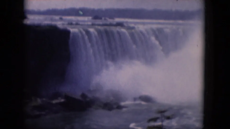1964 8mm frame cataloged to Albany, New York, showing a wide waterfall, rocky foreground, mist, and distant shoreline.