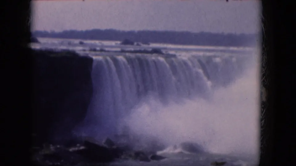 1964 8mm home-movie frame with a broad waterfall, dark cliff edge, spray at the base, and purple-blue archival color.