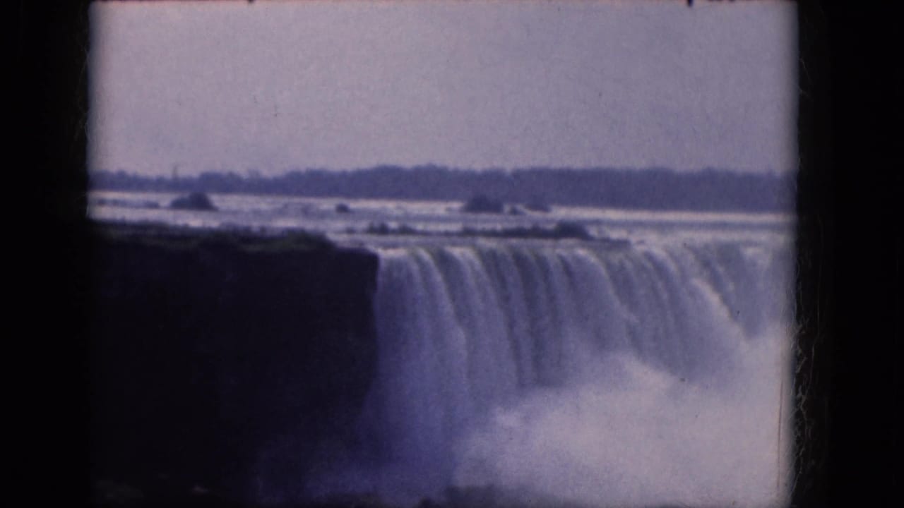 Blue-tinted 8mm frame of Niagara Falls in 1964, showing the waterfall curtain, dark rock edge, spray, and distant tree line.