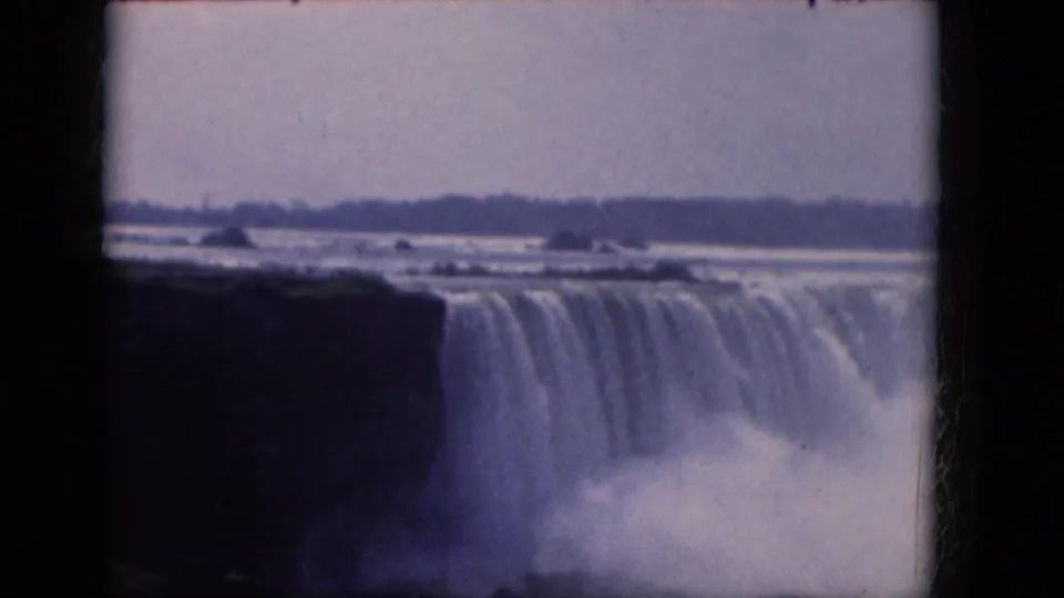 Blue-tinted 8mm frame of Niagara Falls in 1964, showing the waterfall curtain, dark rock edge, spray, and distant tree line.