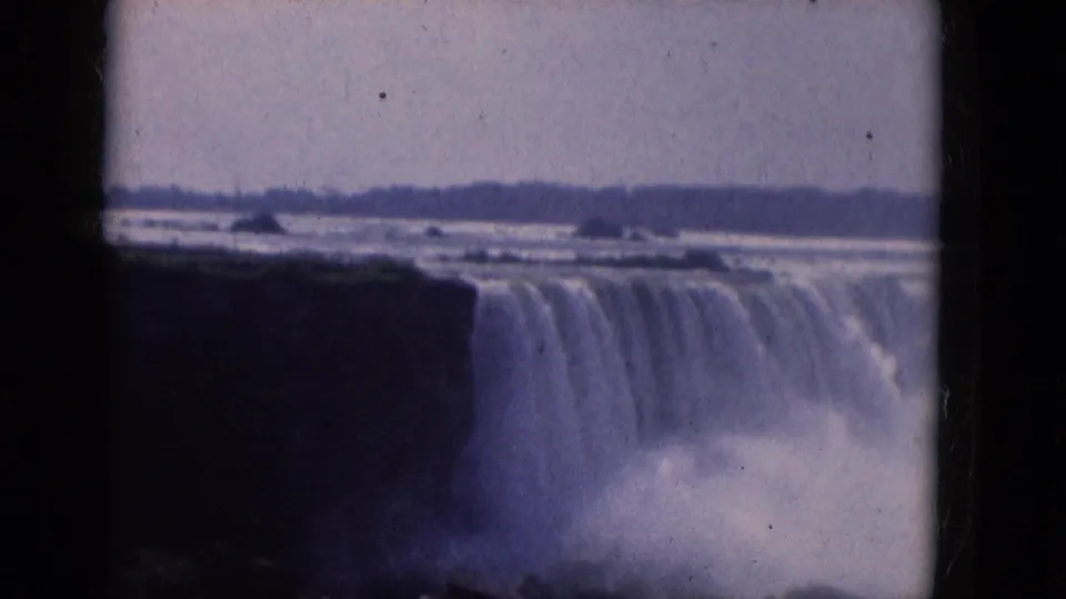 1964 visitor-shot 8mm view of Niagara Falls, with soft focus, purple-blue tint, plunging water, and dense mist below.