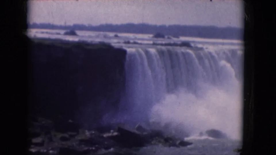 Archival 8mm frame of Niagara Falls in 1964, with water spilling over the rim and spray rising among dark rocks at the base.