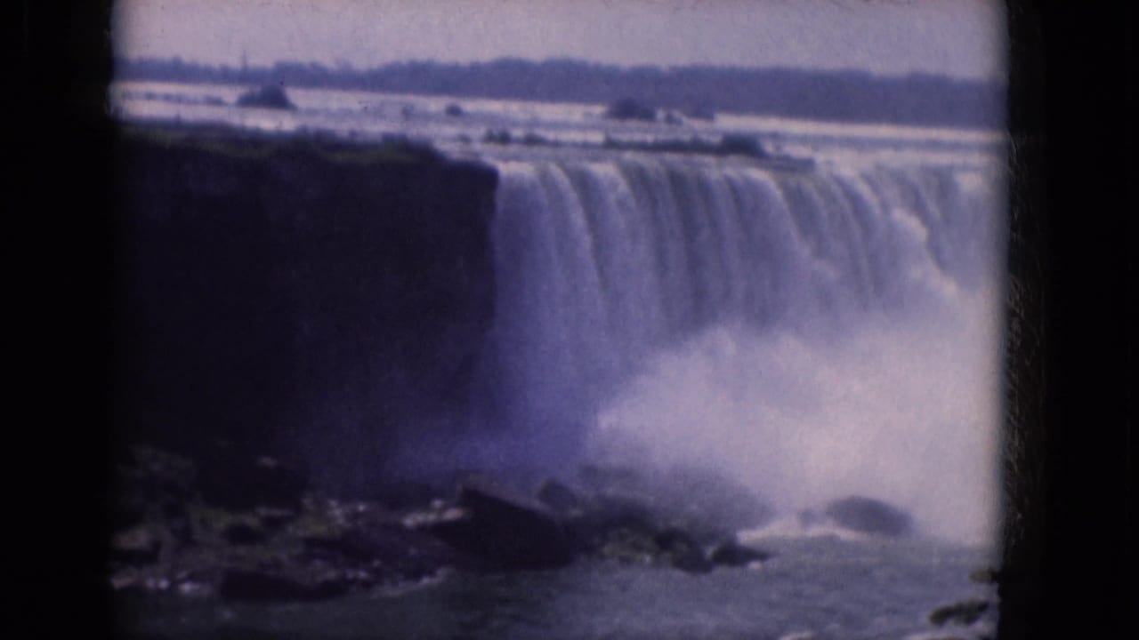 1960s 8mm frame showing a broad waterfall, dark cliff edge, mist plume, and distant shoreline from a visitor vantage point.