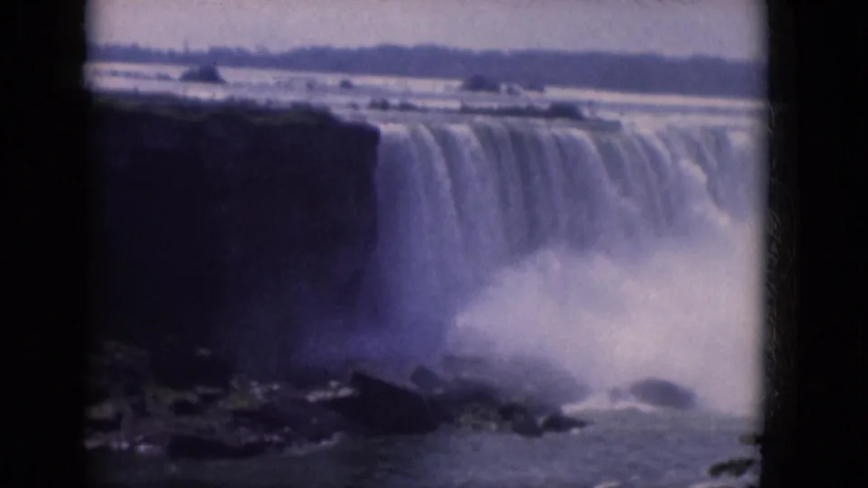 1960s 8mm frame showing a broad waterfall, dark cliff edge, mist plume, and distant shoreline from a visitor vantage point.