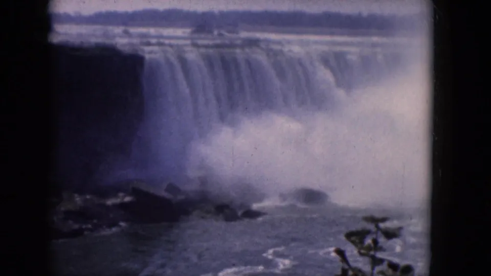 Soft 8mm frame showing cascading water over a cliff, heavy spray, dark rock formations, and churning river water.