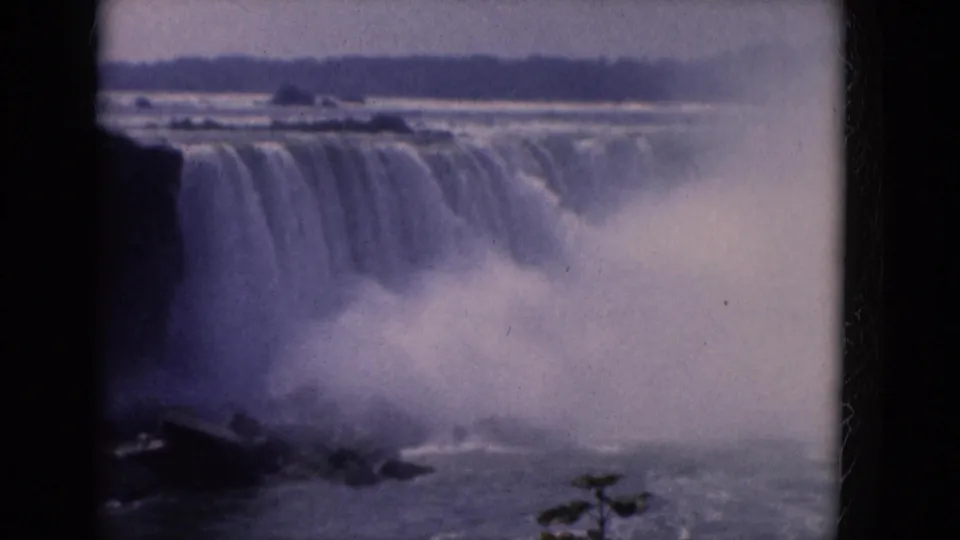 Final 8mm frame with a waterfall receding into mist, dark cliff at left, river below, and faint foreground foliage.