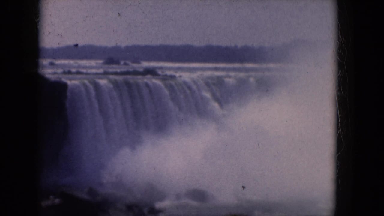 1960s 8mm view of Niagara Falls from the Canadian side, with the waterfall brink, heavy mist, and distant shoreline visible.