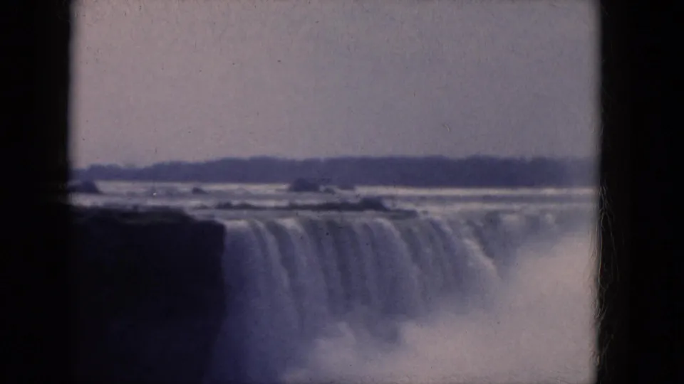Faded 1960s 8mm frame of Niagara Falls, with soft focus, mist at the base, and a low horizon beyond the river.