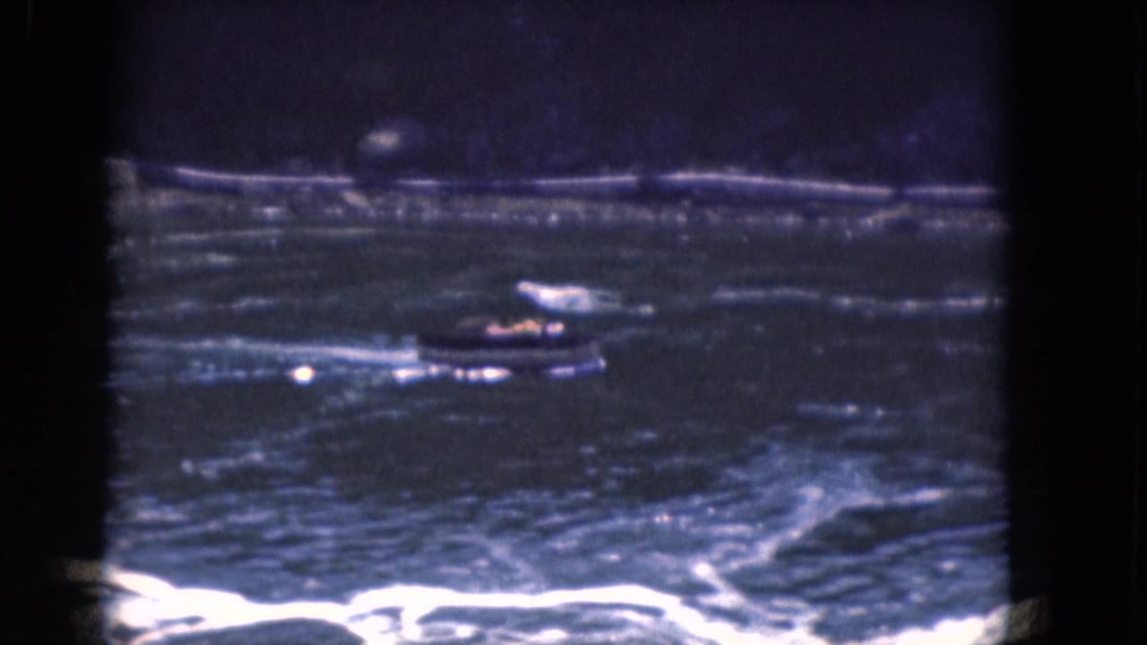 Small motorboat moves across choppy blue water with a white wake and dark shoreline behind it.