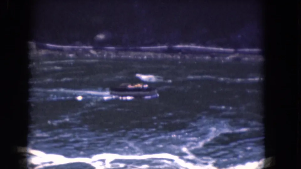 1964 Albany-area 8mm frame showing a small motorboat crossing rough water with a low shoreline in the background.
