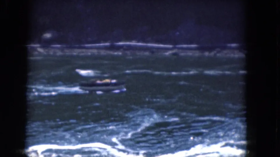 1964 Albany-area 8mm still with a small motorboat crossing dark, textured water beneath a faint shoreline.