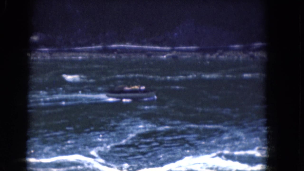 1964 Albany-area 8mm frame showing a small motorboat amid rippled blue water, white foam, and a dark horizontal shoreline.