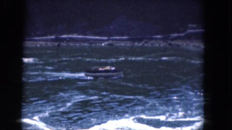 1964 Albany, New York home-movie frame showing a small motorboat moving across rough river water near a distant wooded shore.