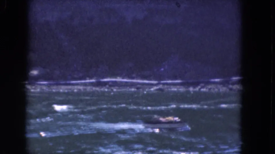 1964 Albany, New York 8mm frame with a small motorboat and wake on a wide river below a soft, dark shoreline.