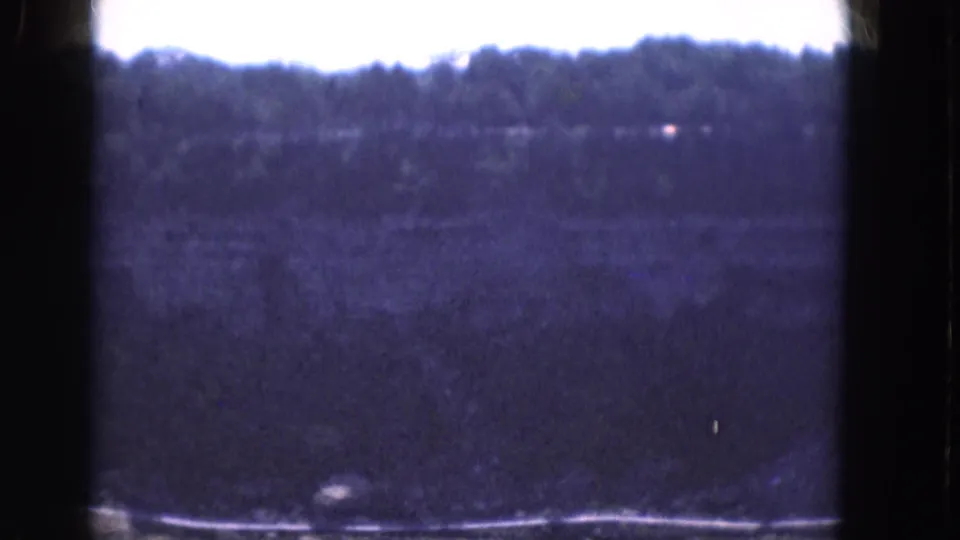 1964 Albany home-movie frame showing a faint small boat on a wide river, with wooded shoreline and bright sky beyond.